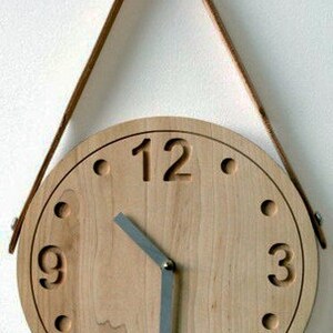 May include: A wooden clock with a leather strap for hanging. The clock face is made of light wood with black numbers and a silver hour and minute hand.