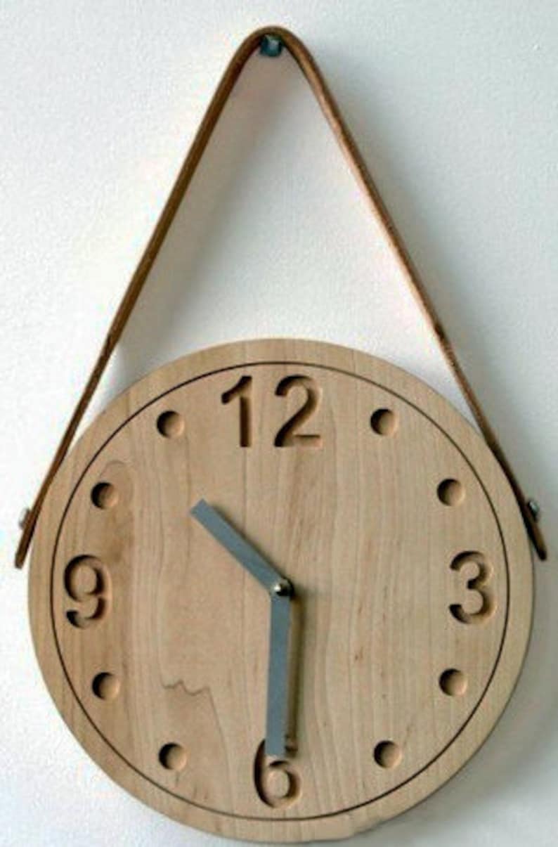 May include: A wooden clock with a leather strap for hanging. The clock face is made of light wood with black numbers and a silver hour and minute hand.