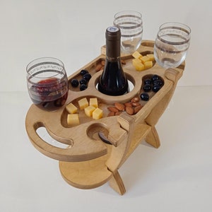 Puede incluir: Bandeja de servicio de madera para vino y queso con una botella de vino tinto, cuatro vasos, cubos de queso, aceitunas y almendras. La bandeja tiene un soporte para botellas integrado y cuatro soportes para vasos.