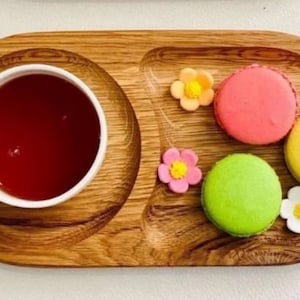 Pode incluir: Uma bandeja de servir de madeira com três compartimentos. A bandeja tem uma chávena de chá, três macarons coloridos e flores decorativas.