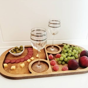 May include: A wooden serving tray with divided sections for snacks and drinks. The tray features a curved shape and is filled with various snacks, including salami, cheese, olives, grapes, and candy. Two wine glasses are placed on the tray.
