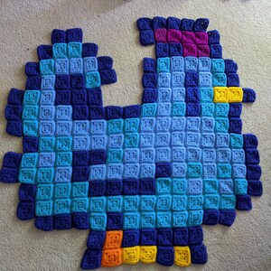 May include: A blue, teal, purple, yellow, and orange crocheted blanket in the shape of a bird. The blanket is made of squares and has a textured surface.