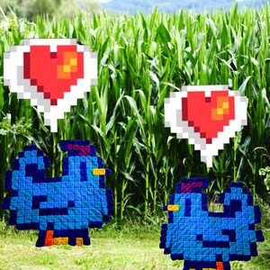 May include: Two blue crocheted birds with red pixelated hearts above them. The birds are in a field of green corn stalks.