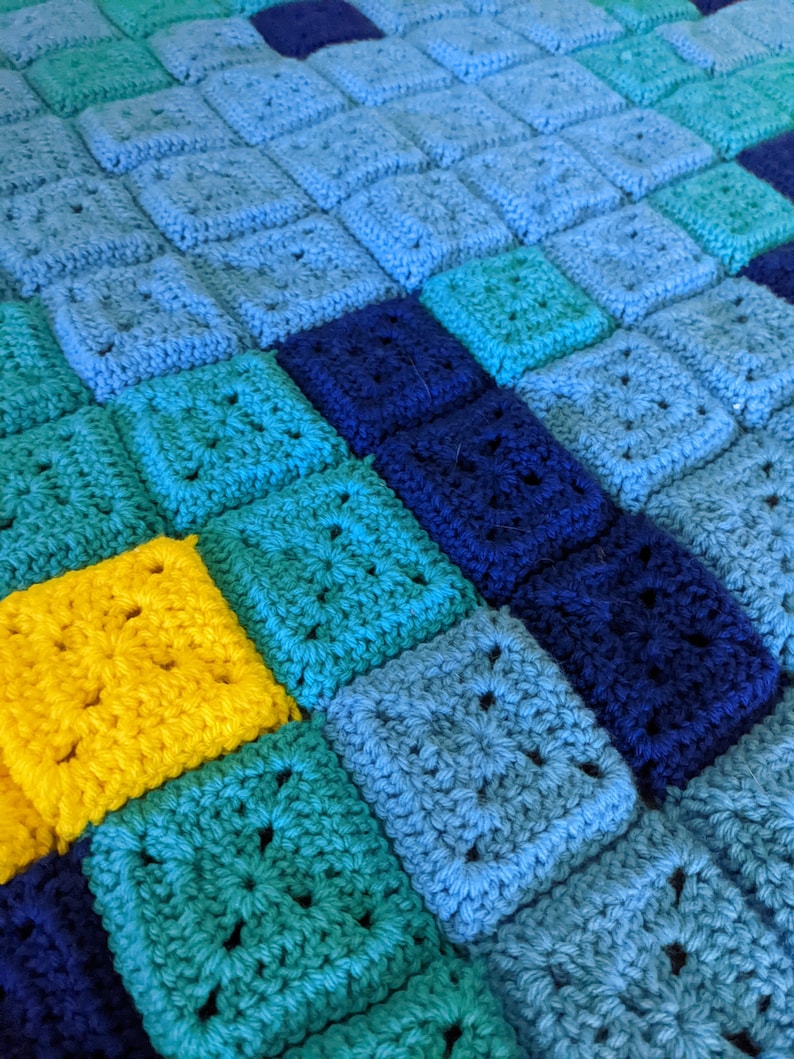 May include: A crocheted blanket with squares in shades of blue, teal, and yellow. The squares are arranged in a diagonal pattern.