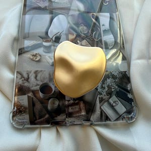May include: Clear phone case with a collage design featuring coffee, books, and perfume. A gold-colored, abstract-shaped phone grip is attached to the case. The case is on a white fabric surface.
