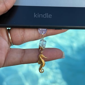 May include: A gold seahorse charm hangs from a Kindle e-reader. The charm is attached to a small, light purple bead and a cluster of clear, blue-tinted beads. The word "Kindle" is printed on the e-reader.