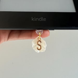 Puede incluir: Un marcapáginas de concha blanca con un dije de letra "S" dorada unido a un lector electrónico Kindle.