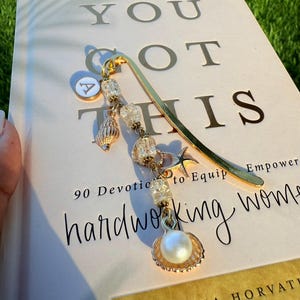 Puede incluir: Un marcapáginas dorado con un dije con una perla, conchas marinas y cuentas transparentes. El marcapáginas está sobre un libro con las palabras "YOU GOT THIS" y "hardworking women" impresas en la portada.
