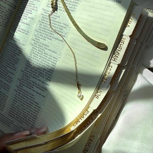 Könnte beinhalten: Ein goldfarbener Metall-Lesezeichen mit einem Kreuzanhänger und einer kleinen Perle, das auf den offenen Seiten eines Buches liegt. Das Buch hat beschriftete Register und Text. Das Lesezeichen ist ein dekoratives Accessoire.