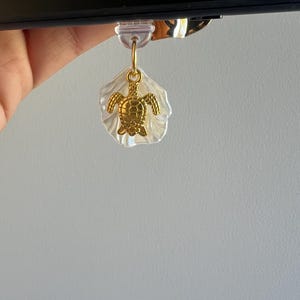 May include: A gold turtle charm attached to a clear plastic shell bookmark. The bookmark is attached to a black Kindle e-reader.