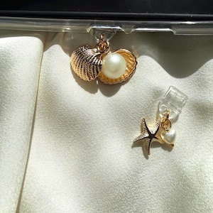 May include: Gold-toned seashell and starfish charms with pearl accents. The seashell charm features a pearl nestled inside. The starfish charm has a small pearl and a heart-shaped accent. The charms are attached to a clear plastic clip.