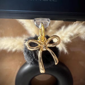May include: A black faux fur pom pom with a gold bow attached to a clear phone ring holder. The ring holder is attached to a black Kindle e-reader.