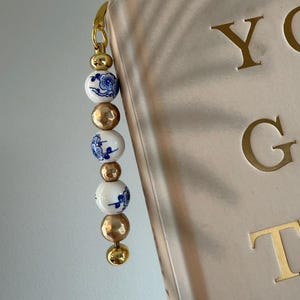 Op de afbeelding: Een decoratieve bladwijzer met een goudkleurige sluiting en kralen, afgewisseld met witte kralen versierd met blauwe bloemmotieven. De bladwijzer is bevestigd aan een boek met gouden letters.
