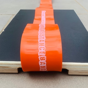 May include: An orange resistance band with white text reading "RESISTANCE BAND" is laid across a black textured surface. The surface is on a wooden platform. The band is likely used for exercise and fitness.
