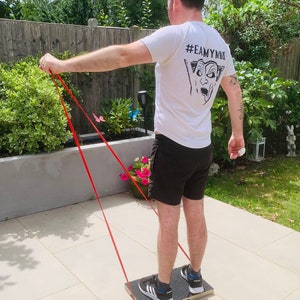 May include: A person exercising outdoors using a resistance band and a wooden balance board. The person is wearing a white t-shirt with a graphic and black shorts. The resistance band is red.
