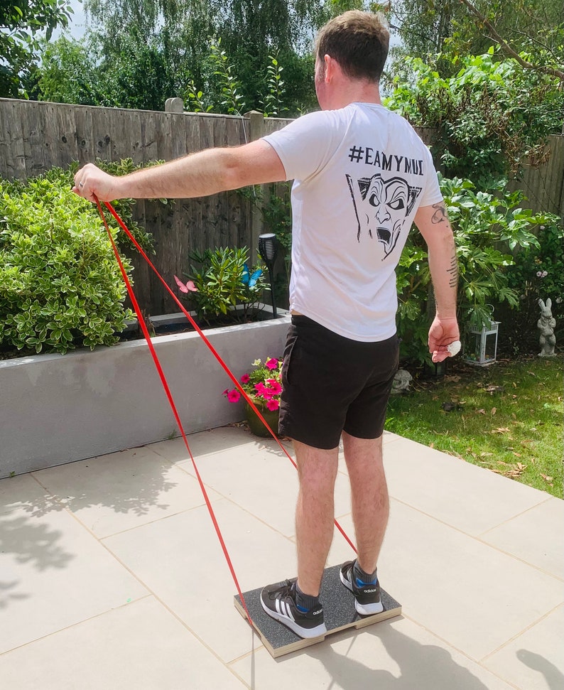 May include: A person exercising outdoors using a resistance band and a wooden balance board. The person is wearing a white t-shirt with a graphic and black shorts. The resistance band is red.