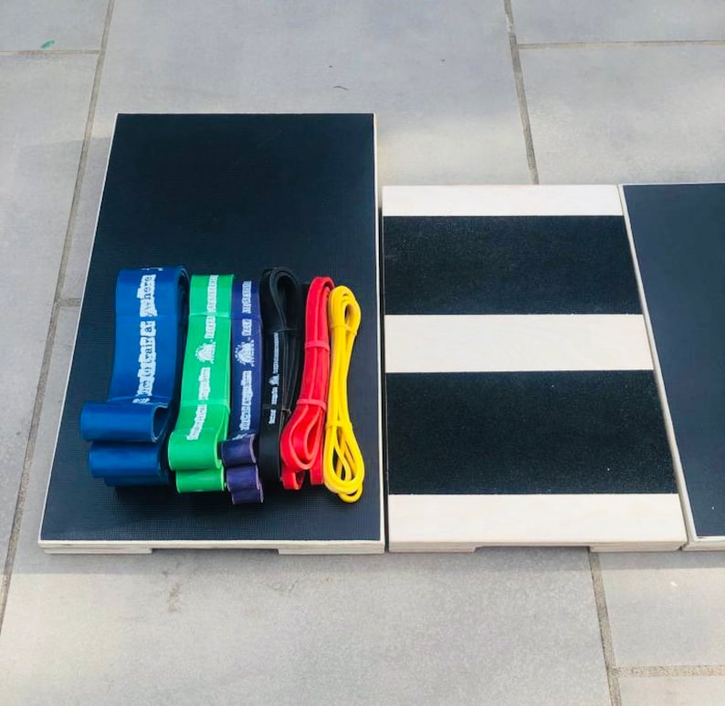 May include: A collection of resistance bands in blue, green, purple, black, red, and yellow, arranged on a black wooden platform. The bands are of varying thicknesses and lengths. Adjacent to the platform is a wooden step with black and white stripes.