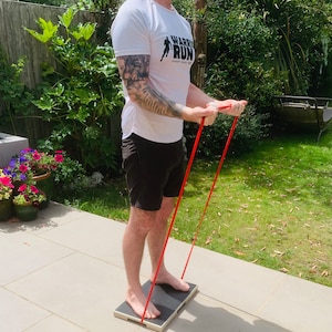 May include: A person is exercising with a red resistance band, standing on a black and white platform. The person is wearing a white t-shirt with the text "WARRIOR RUN" and black shorts. The background includes green foliage and a sunny outdoor setting.