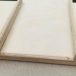 May include: Close-up of a light-colored wooden object with a rectangular shape. The object has a layered construction, with visible edges and a smooth, flat surface. Black square pads are affixed to the corners. The object appears to be a part of a larger structure.