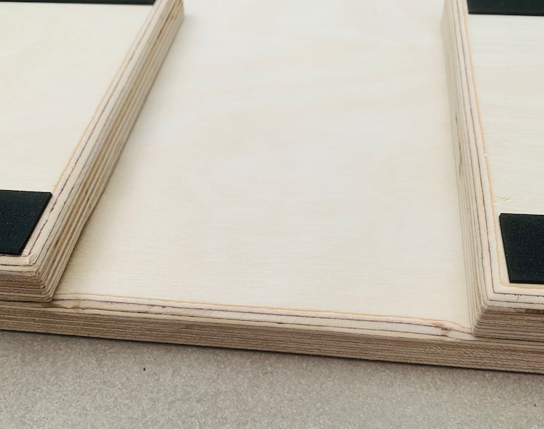 May include: Close-up of a light-colored wooden object with a rectangular shape. The object has a layered construction, with visible edges and a smooth, flat surface. Black square pads are affixed to the corners. The object appears to be a part of a larger structure.