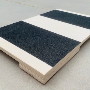 May include: A wooden balance board with alternating black and white stripes. The board has a rectangular shape with rounded corners and small feet on the bottom. The black stripes have a textured, non-slip surface.