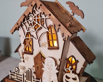 Pode incluir: Uma casa de Halloween de madeira com uma janela iluminada, um vampiro, uma abóbora e um morcego no telhado. A casa tem uma teia de aranha na frente e uma cerca em torno da base.