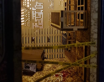 May include: A miniature wooden model of a city alleyway with a crime scene marked off with yellow tape. The scene includes a chalk outline of a body, a wooden fence, and a building with a balcony.