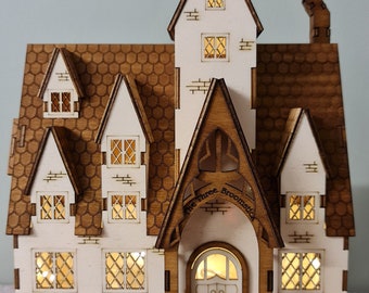 Puede incluir: Un modelo de madera de una casa con un techo puntiagudo y un letrero que dice "The Three Broomsticks". La casa tiene un exterior blanco con molduras marrones y ventanas. El modelo está iluminado con pequeñas luces. La casa está sentada sobre un libro rojo con letras doradas que dicen "Jules Verne Seven Novels".