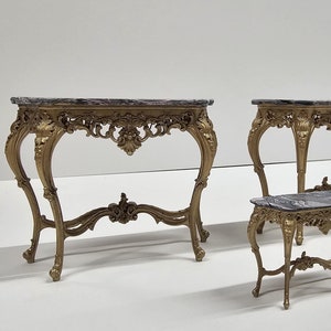 May include: Four ornate gold-painted console tables with marble tops. The tables feature intricate carvings and scrolled legs. One table is smaller than the others.