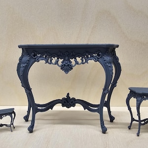 Peut inclure: Trois tables en bois de couleur gris foncé, ornées de sculptures complexes. La plus grande table est une console avec un plateau rectangulaire et des pieds courbes. Les deux plus petites tables sont des tables d'appoint avec des plateaux carrés et des pieds courbes.
