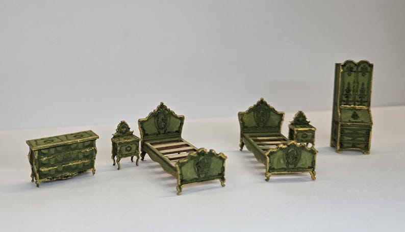 May include: A miniature bedroom set in a vintage style. The set includes a chest of drawers, two bedside tables, two beds, and a wardrobe. The furniture is painted dark green with gold accents, creating an antique aesthetic.