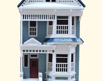 May include: A miniature model of a blue and white Victorian house with a white picket fence and a red door. The house has a bay window and a garage door. The model is sitting on a book.