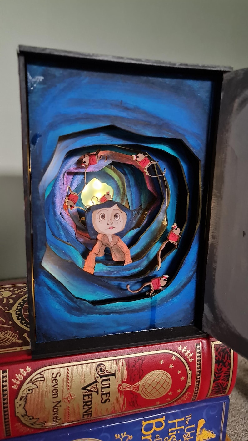 Pu&ograve; includere: Un'opera d'arte di libro pop-up 3D con una ragazza con gli occhi azzurri e un vestito rosso in piedi in un tunnel blu e oro vorticoso. Il tunnel &egrave; circondato da piccole figure rosse con le code.