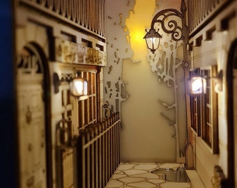 May include: A miniature wooden model of a city alleyway with a street lamp, a building with a balcony, and a cobblestone pathway. The model is lit with warm white LED lights.
