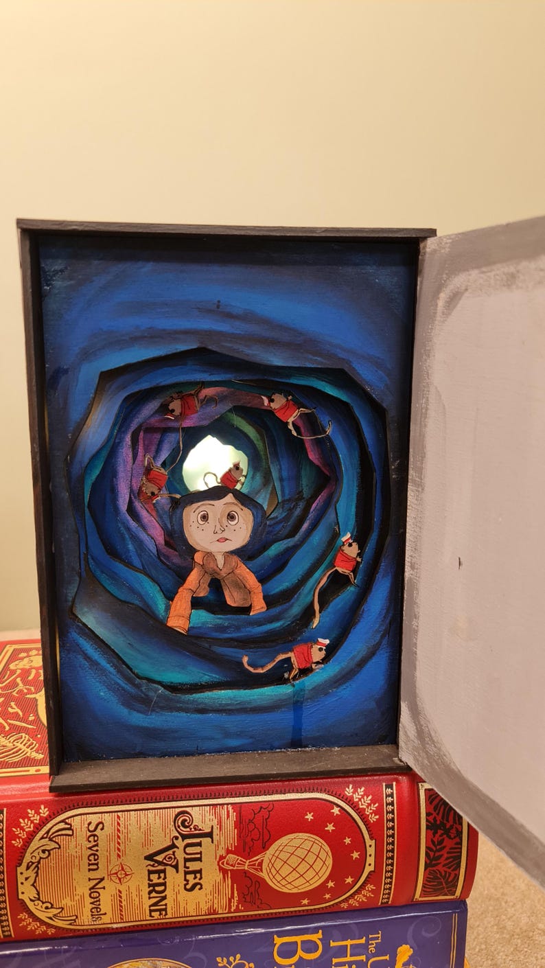 Pu&ograve; includere: Un diorama dipinto a mano di una ragazza con i capelli blu in piedi in un tunnel blu vorticoso con topi rossi e bianchi che strisciano intorno a lei. Il diorama &egrave; incorniciato in una cornice nera e si trova sopra una pila di libri.