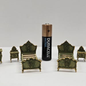 May include: A miniature bedroom furniture set in a vintage style. The set includes a chest of drawers, two bedside tables, two beds, and a wardrobe, all in green and gold. A Duracell Ultra battery is included for scale.