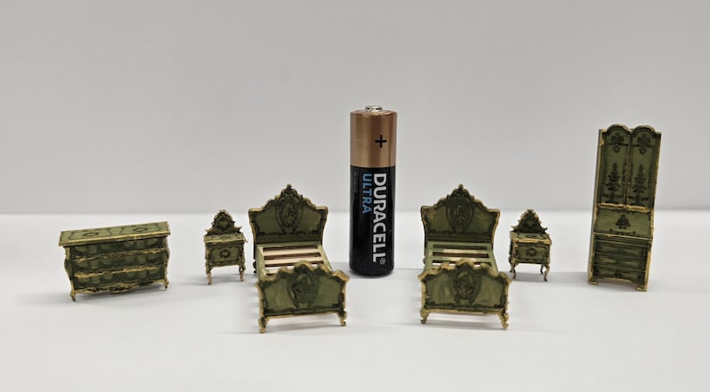 May include: A miniature bedroom furniture set in a vintage style. The set includes a chest of drawers, two bedside tables, two beds, and a wardrobe, all in green and gold. A Duracell Ultra battery is included for scale.