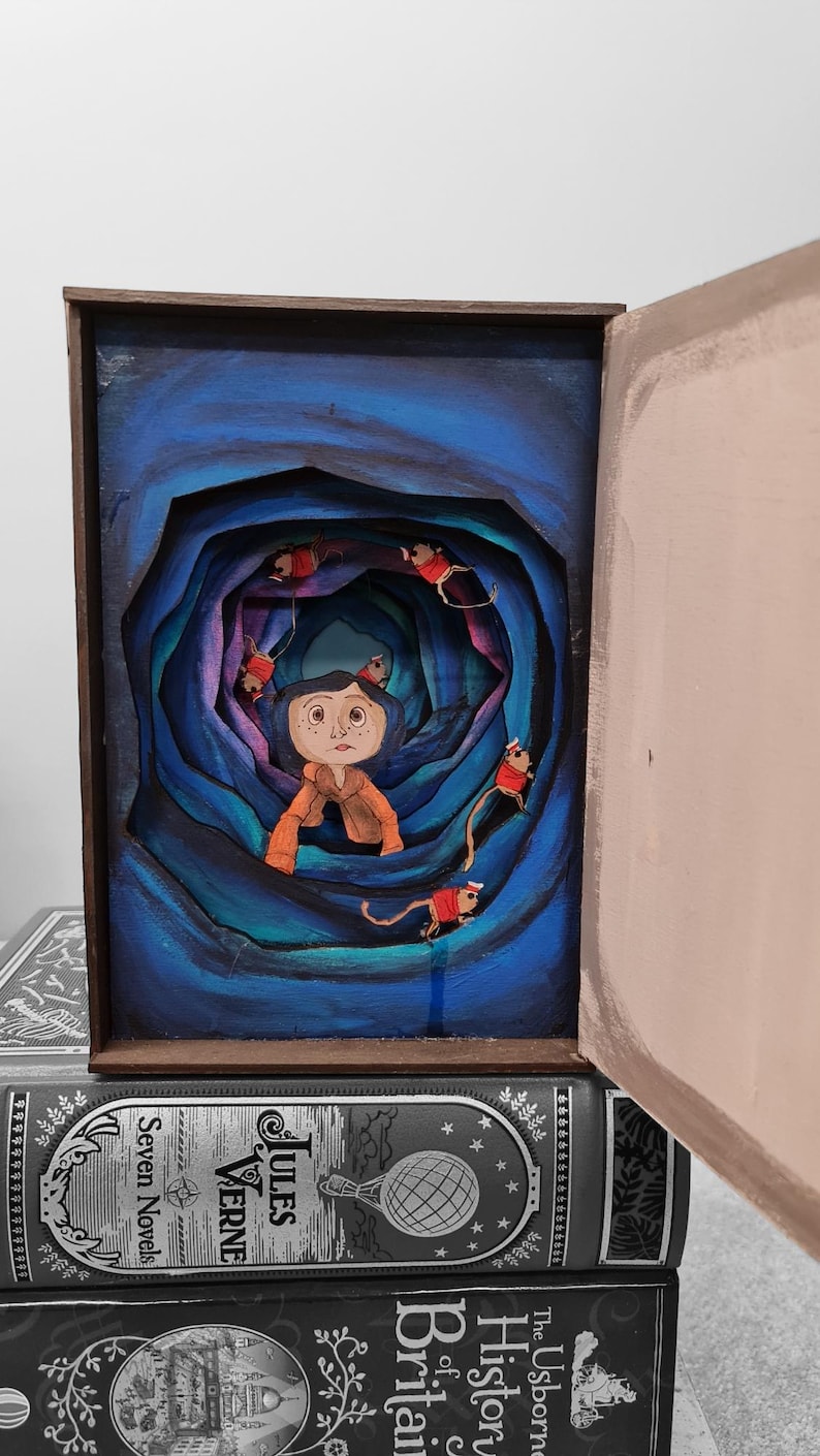 Pu&ograve; includere: Una scatola di legno dipinta a mano con un diorama 3D all'interno. Il diorama rappresenta una ragazza con i capelli blu e un vestito rosso in piedi in un tunnel blu e viola vorticoso. Ci sono diversi piccoli topi rossi con code bianche che pendono dalle pareti del tunnel.