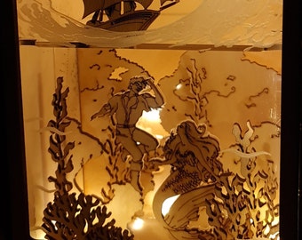 Puede incluir: Una escena luminosa de madera con un barco navegando en una ola, una sirena y un hombre en el agua, y coral y algas marinas. La escena está iluminada desde atrás, creando un brillo cálido.