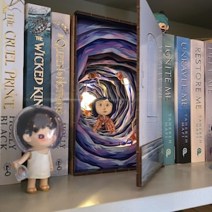 May include: A shelf displays a diorama of a tunnel scene with a figure, lit by a small bulb. Beside it are several books, including "The Cruel Prince" and "Ignite Me." A small figurine stands on the shelf.