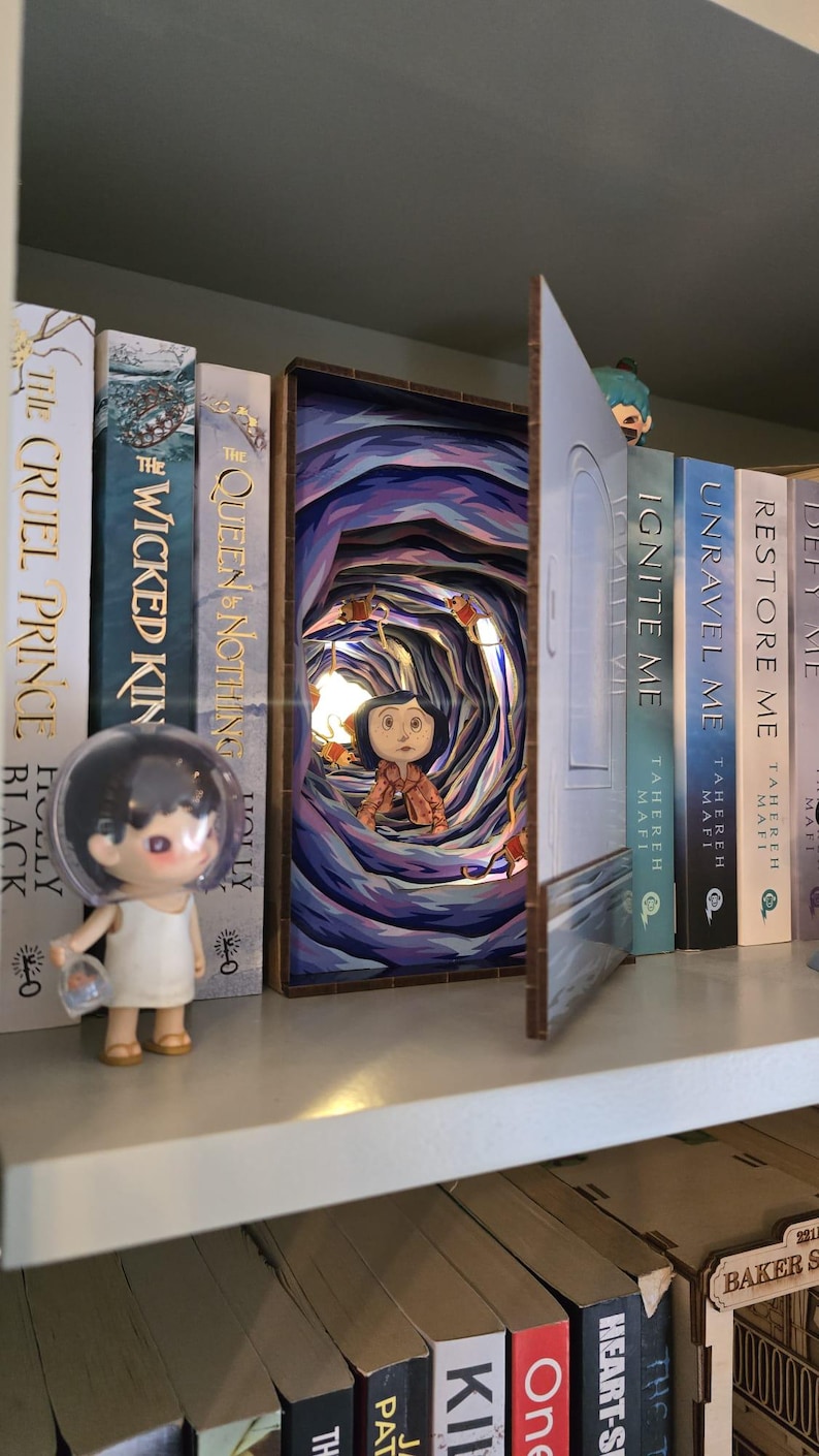 May include: A shelf displays a diorama of a tunnel scene with a figure, lit by a small bulb. Beside it are several books, including "The Cruel Prince" and "Ignite Me." A small figurine stands on the shelf.