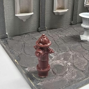 Op de afbeelding: Een miniatuur, bordeauxrode brandkraan staat op een grijs geplaveid oppervlak, onderdeel van een diorama. De achtergrond toont een grijze muur met decoratieve nissen en pilaren. De scène roept stedelijke architectuur op.