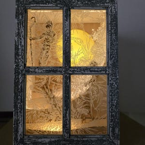 Peut inclure: Un cadre de fenêtre en bois avec un panneau de verre givré. Le verre est gravé d'un motif d'arbre et de lune. La fenêtre est éclairée par derrière, créant une lueur chaude.