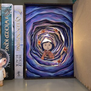 May include: A layered art piece depicting a character in a tunnel with a blue and purple color scheme. The artwork is illuminated with small lights and surrounded by books on a shelf. The character is wearing a pink outfit.