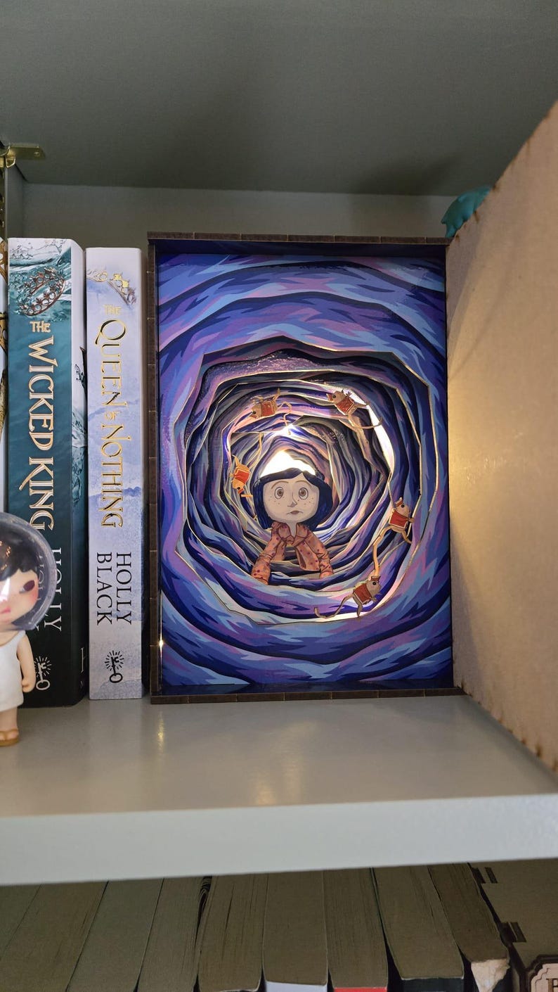 May include: A layered art piece depicting a character in a tunnel with a blue and purple color scheme. The artwork is illuminated with small lights and surrounded by books on a shelf. The character is wearing a pink outfit.