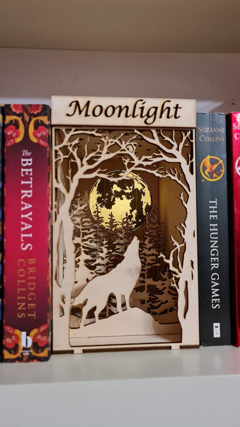 May include: A wooden cutout of a wolf howling at the moon in a forest setting. The cutout is labeled "Moonlight" and is displayed on a bookshelf.