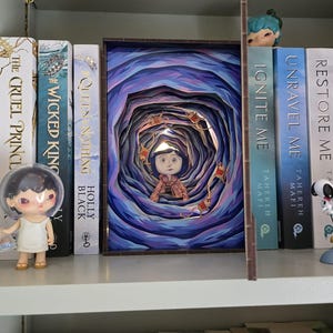 May include: A layered art piece depicting a girl in a tunnel with blue and purple hues. The artwork is displayed on a shelf with books and figurines. The books include titles like "The Cruel Prince" and "Queen Nothing".