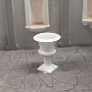 May include: A small, white, decorative urn with a fluted body and a wide rim. The urn sits on a square base. The background features architectural elements, including columns and arched niches, suggesting a classical theme.