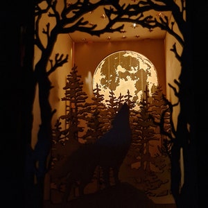 May include: A wooden silhouette of a wolf howling at the moon in a forest setting. The moon is full and bright, and the trees are tall and dark. The scene is lit from behind, creating a warm glow.