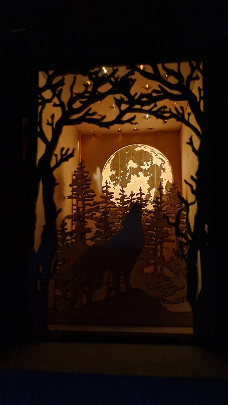 May include: A wooden silhouette of a wolf howling at the moon in a forest setting. The moon is full and bright, and the trees are tall and dark. The scene is lit from behind, creating a warm glow.